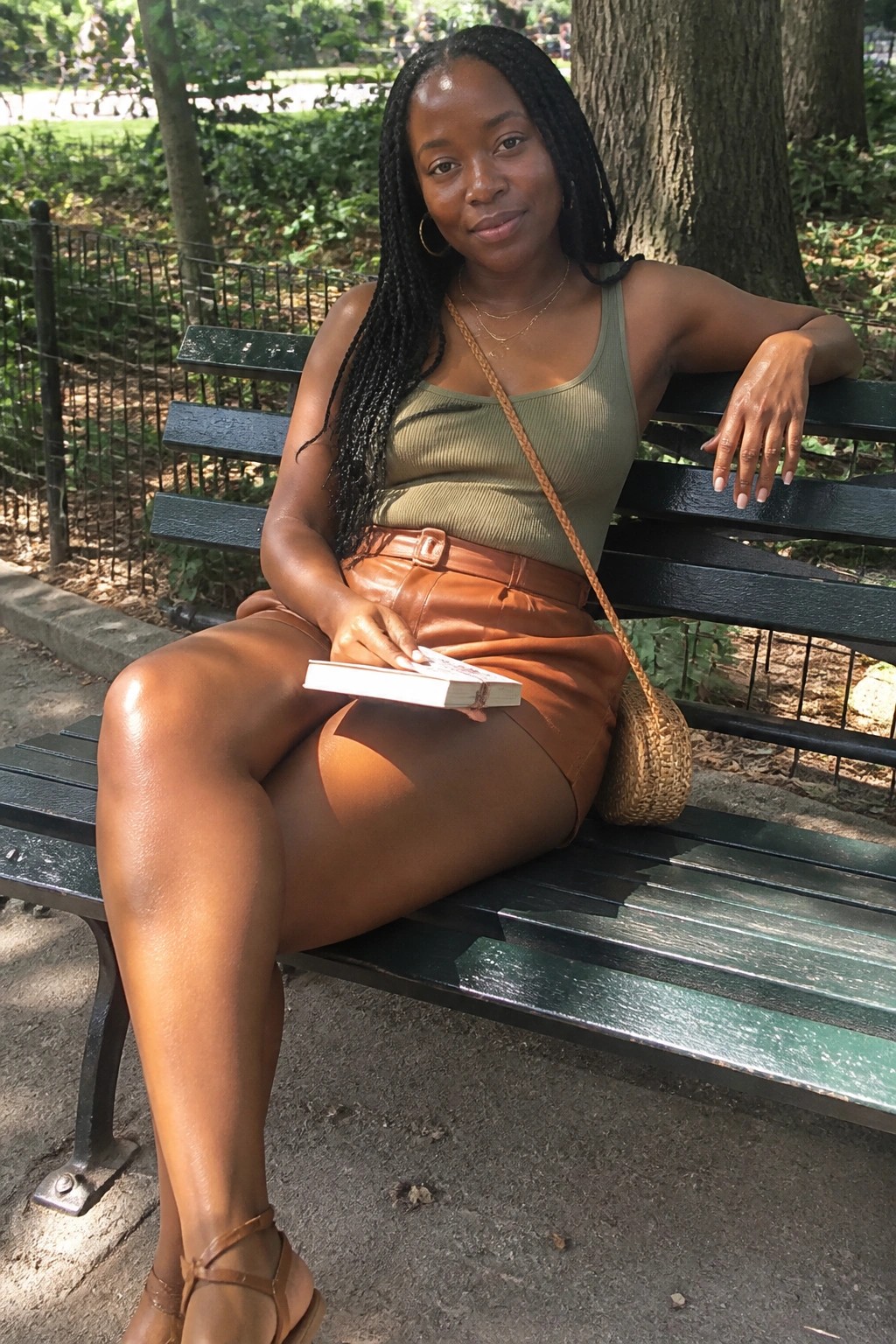 Woman with braids in green tank top and brown shorts on park bench holding book