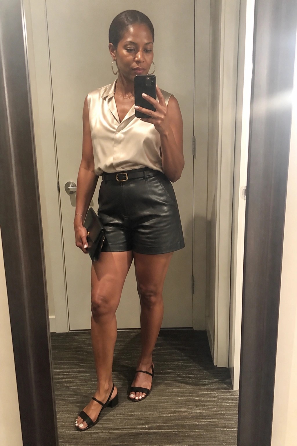 Woman in beige sleeveless blouse and black leather shorts takes mirror selfie.