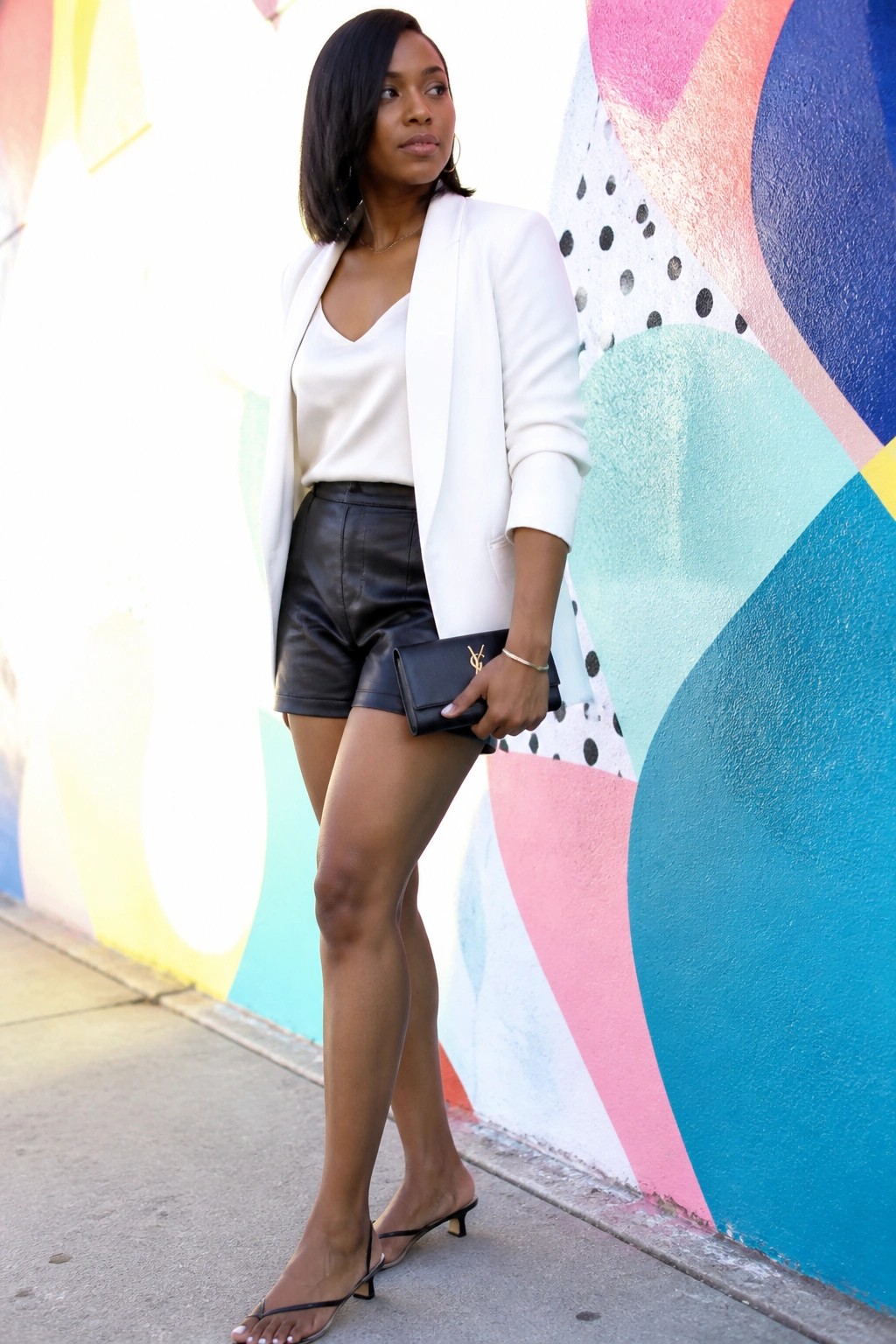 Woman in white blazer and black leather shorts holding YSL clutch by colorful mural