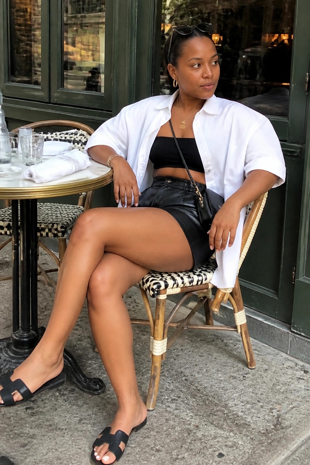 Woman wearing black leather shorts with a black bandeau top, open white oversized button down shirt, black slide sandals, and gold jewelry including a crossbody bag.