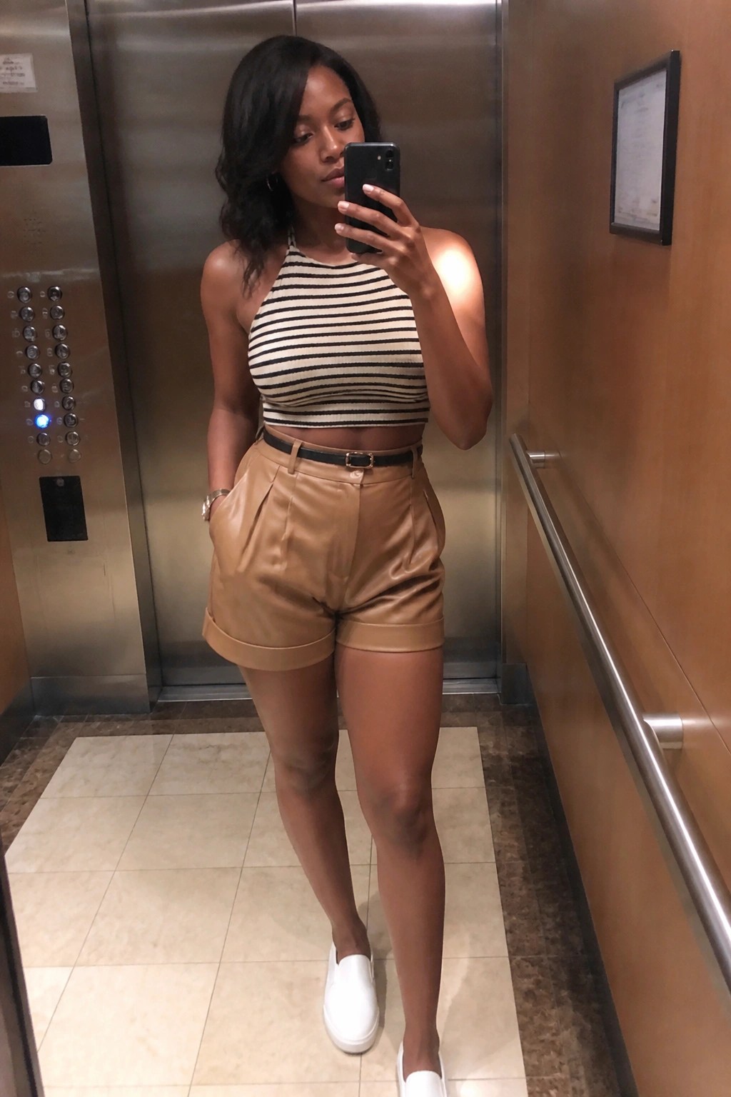 Woman in striped halter crop top and tan shorts taking elevator mirror selfie.