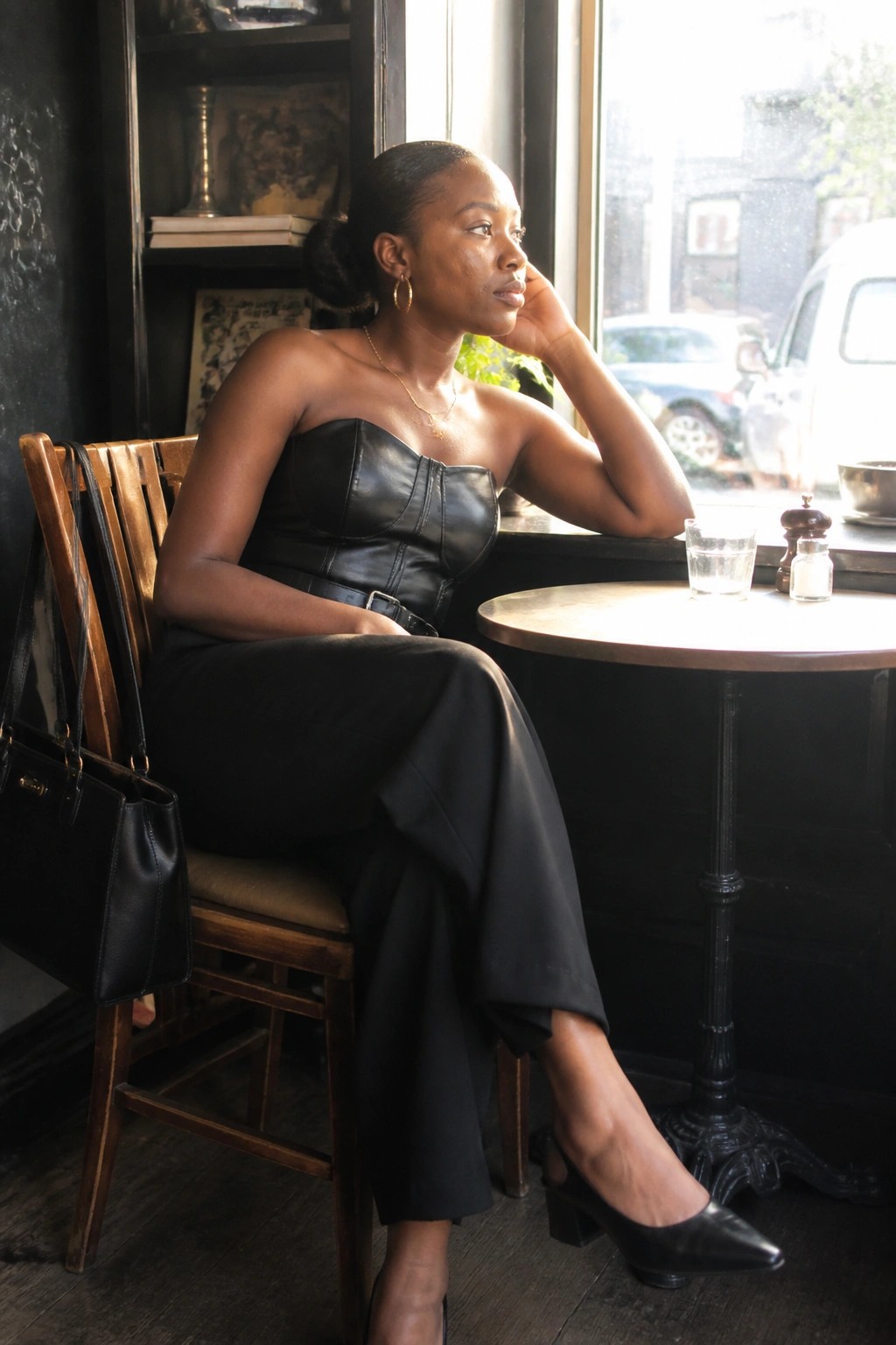 Woman in black leather strapless corset top paired with wide black bottoms black pointed block heel shoes and gold necklace with black handbag nearby