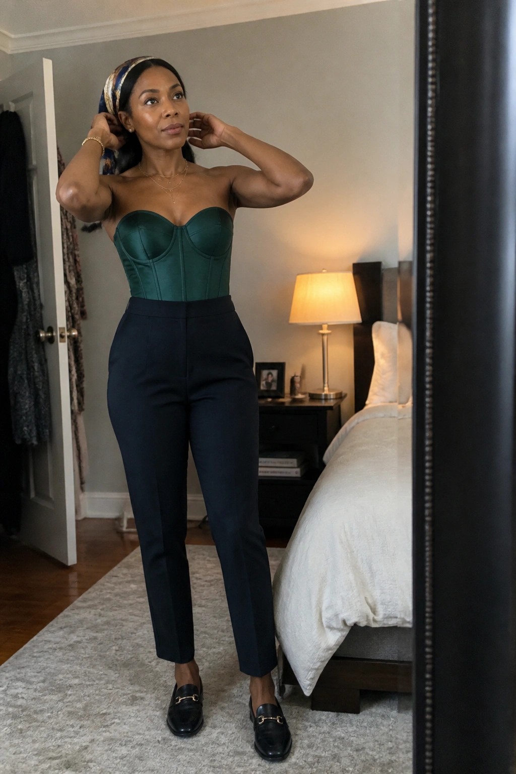 Woman wearing a strapless emerald green satin corset with navy high waisted trousers black loafers and a patterned headwrap standing in a bedroom