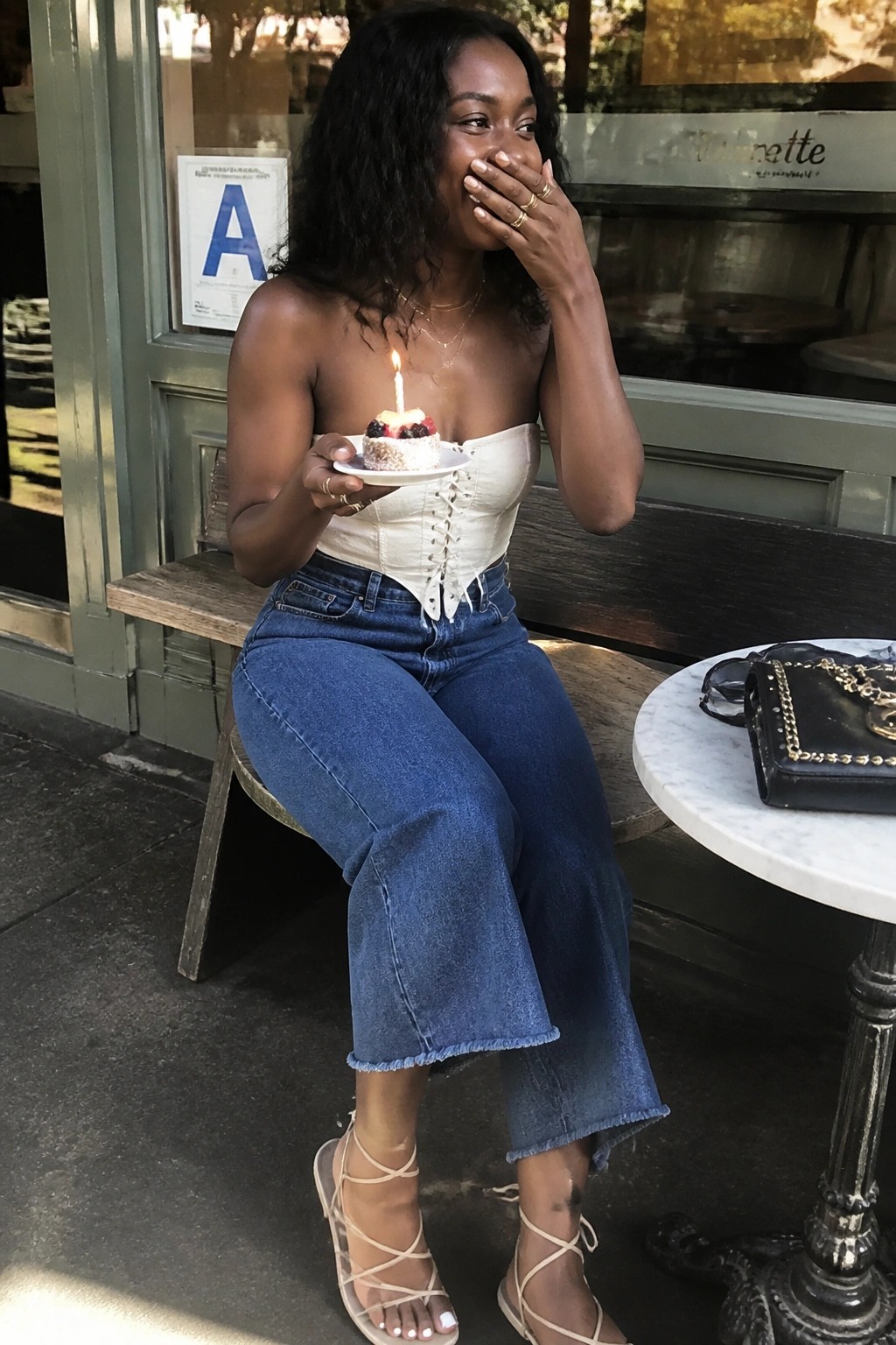 Black woman seated outdoors wearing a white strapless lace-up corset top with blue wide-leg cropped frayed jeans and nude strappy high heels while holding a small birthday cake with a lit candle.