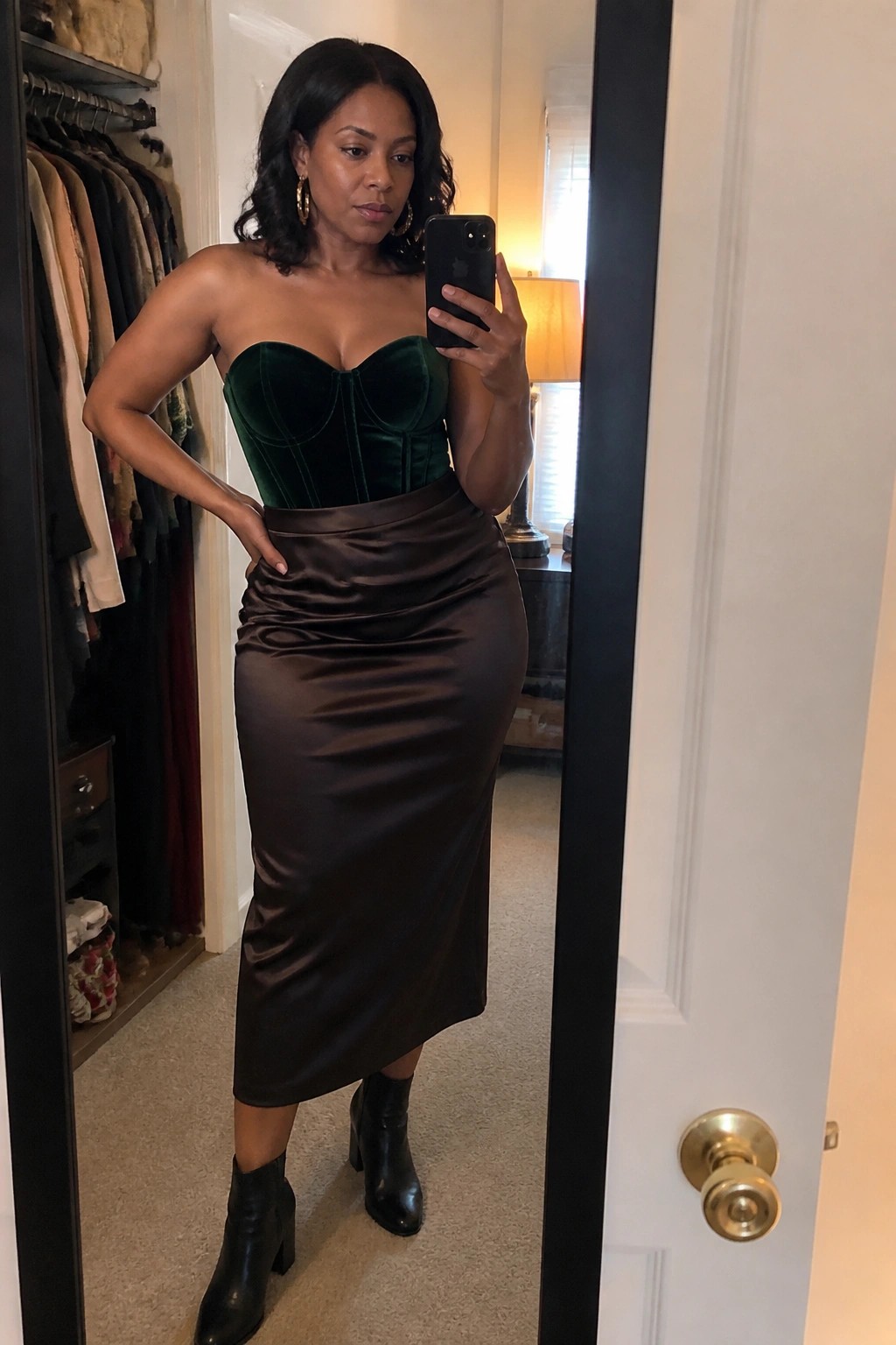 Woman wearing a strapless green velvet corset top with a brown satin midi skirt and black heeled ankle boots while taking a mirror selfie.