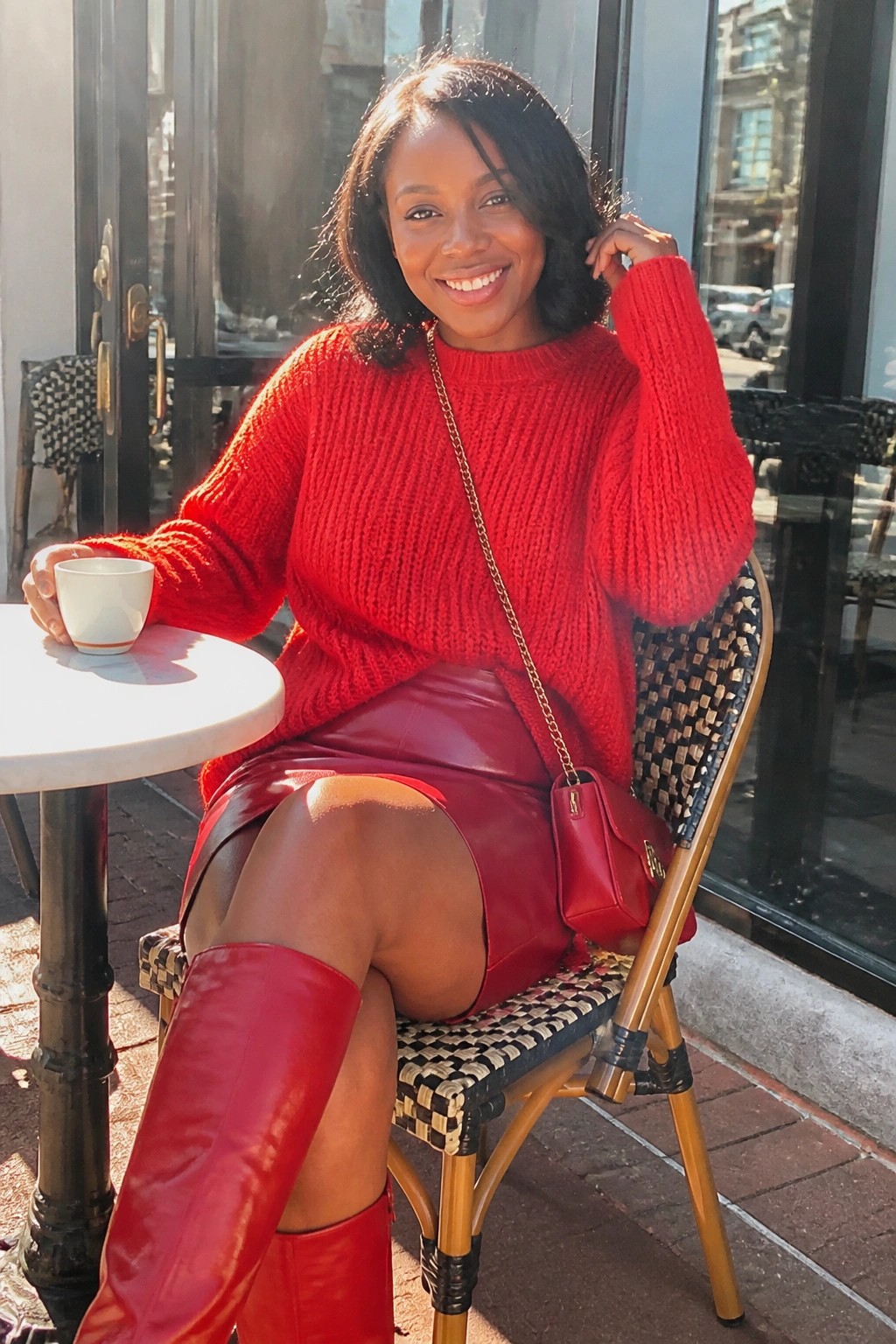 Smiling woman in red knit sweater, leather skirt, and boots at sunny cafe table.