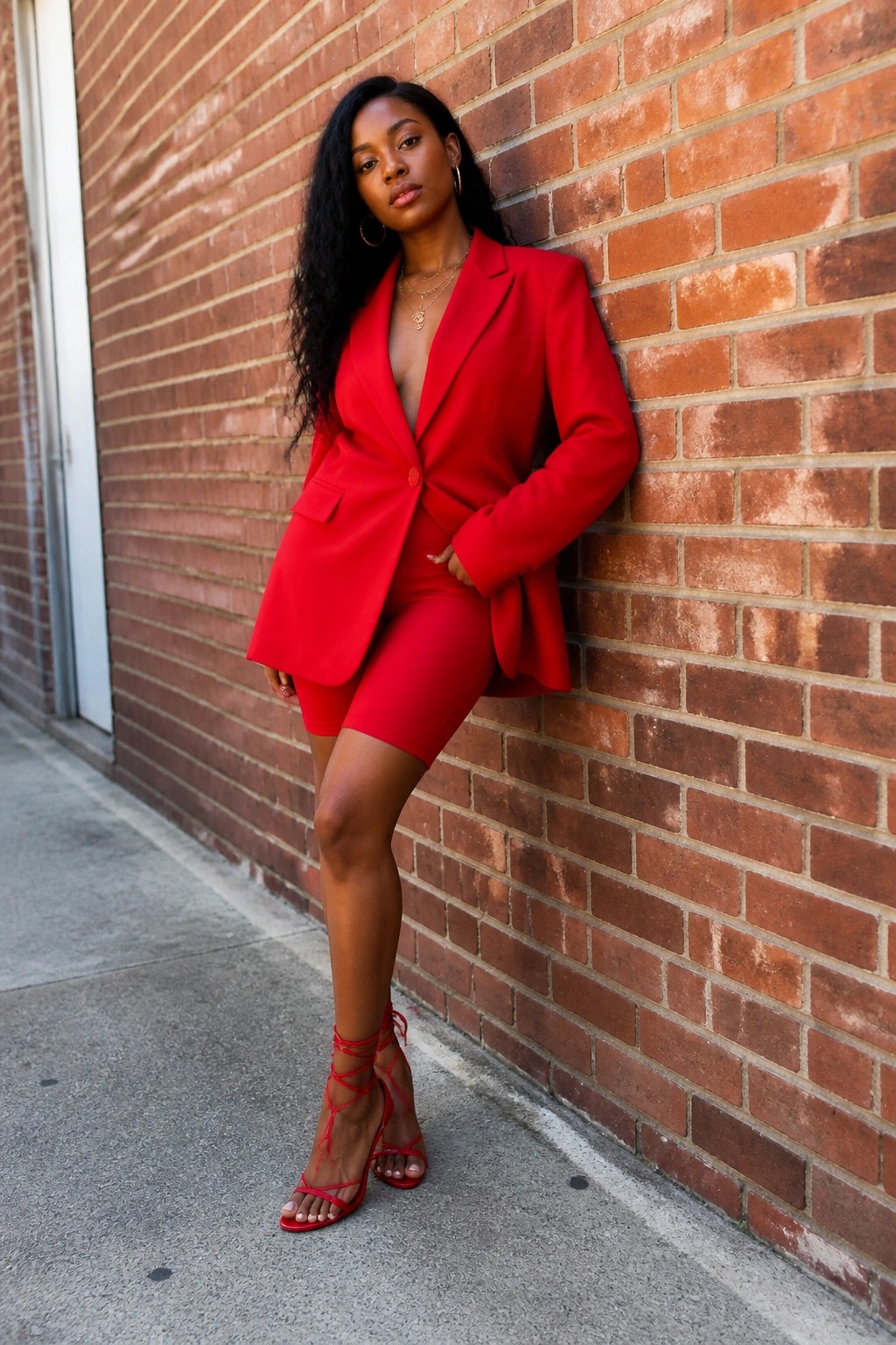 Woman in red blazer and shorts leans against brick wall wearing strappy heels