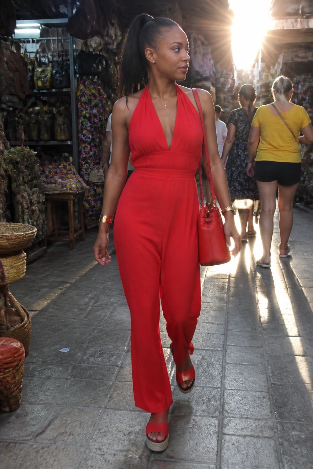 Woman in red halter jumpsuit carrying red bag on sunny market street