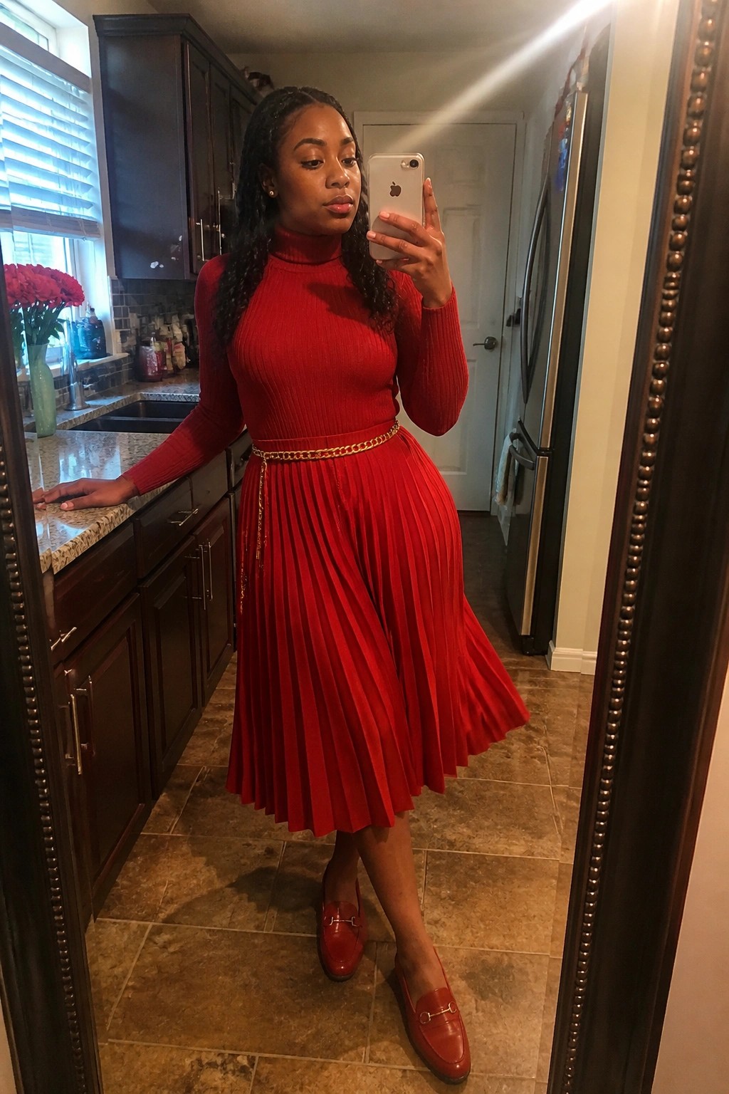 A woman wearing an all-red outfit with a ribbed turtleneck sweater, pleated midi skirt, gold chain belt, and matching red loafers, posing in a kitchen.