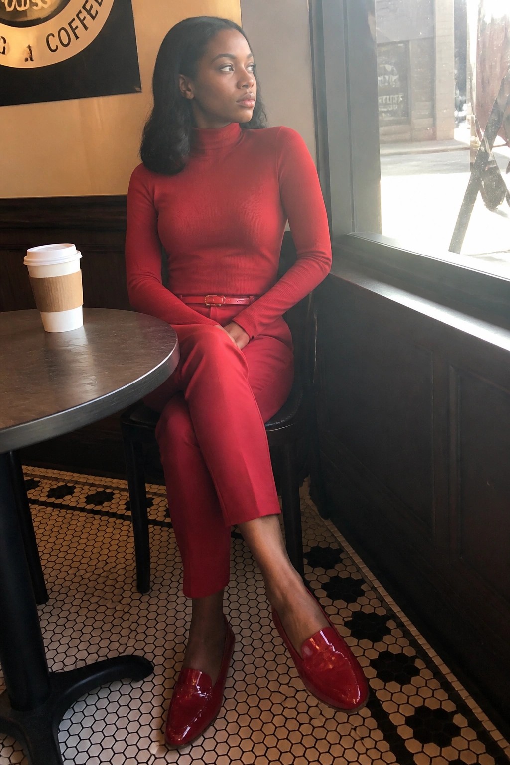 A Black woman in a fitted red turtleneck sweater tucked into red cropped pants with a thin red belt and glossy red patent loafers sitting at a cafe table.