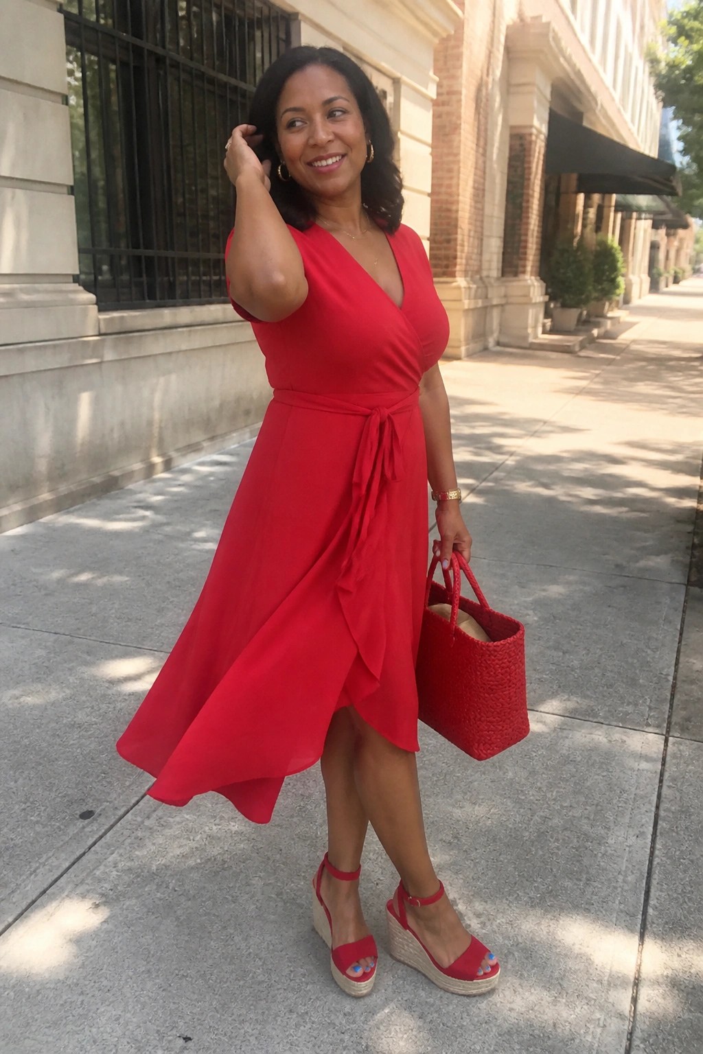 Woman in flowing red wrap dress holding red bag on sunny sidewalk