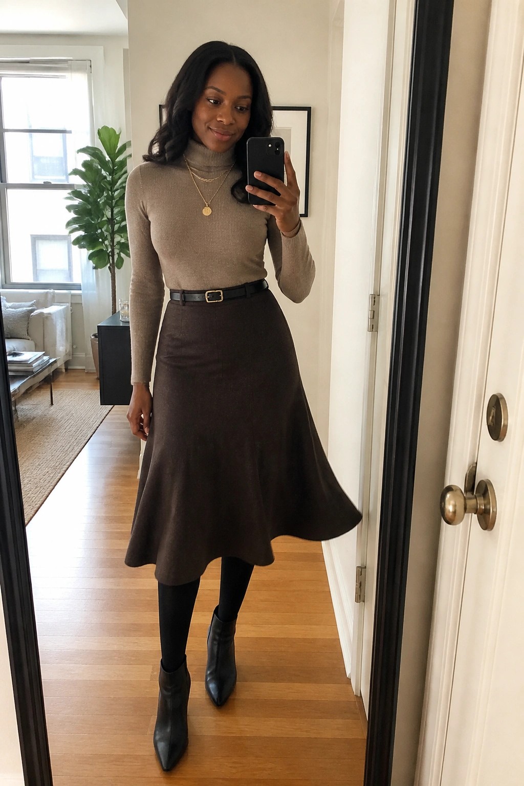 Woman in beige turtleneck and brown flared skirt taking mirror selfie in hallway