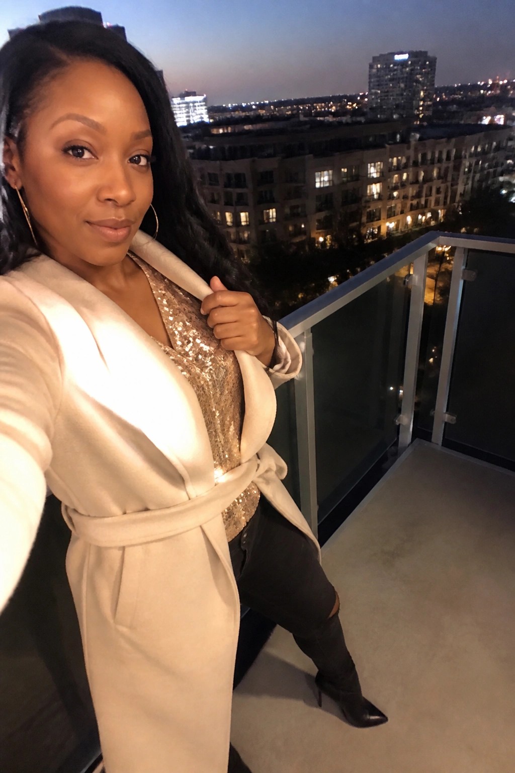 Woman wearing a cream belted coat over a gold sequin top paired with black ripped jeans and black knee high boots.