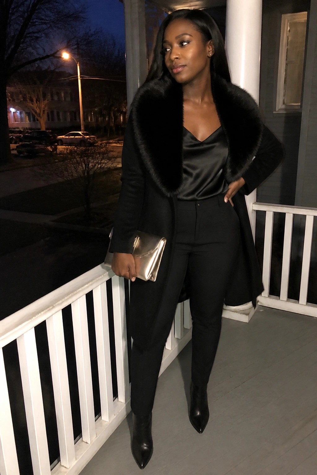 Woman in black fur-collared coat holding gold clutch on porch at night