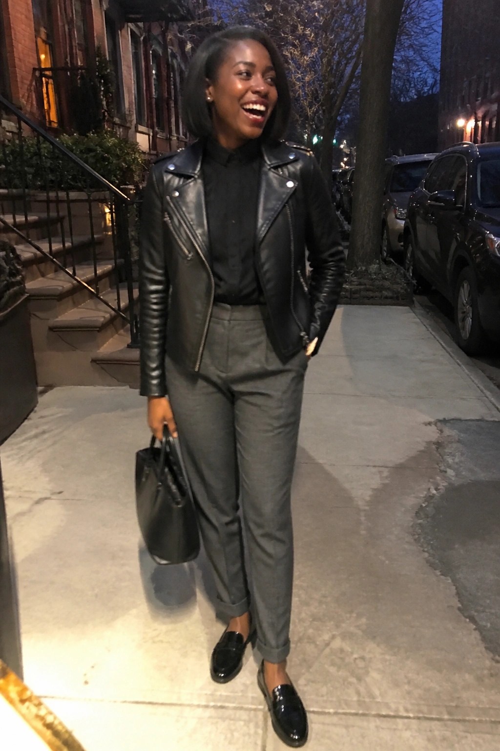 Smiling woman in black leather jacket and gray pants holds bag on city sidewalk