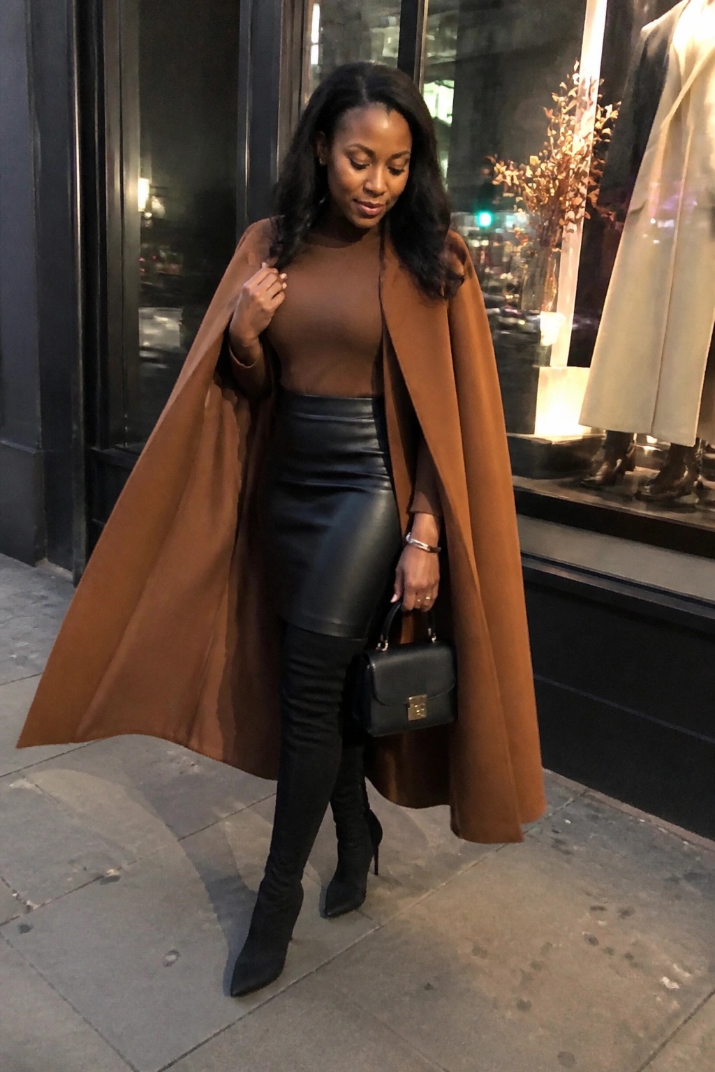 Woman in flowing brown coat, black leather skirt, and thigh-high boots holding handbag at night