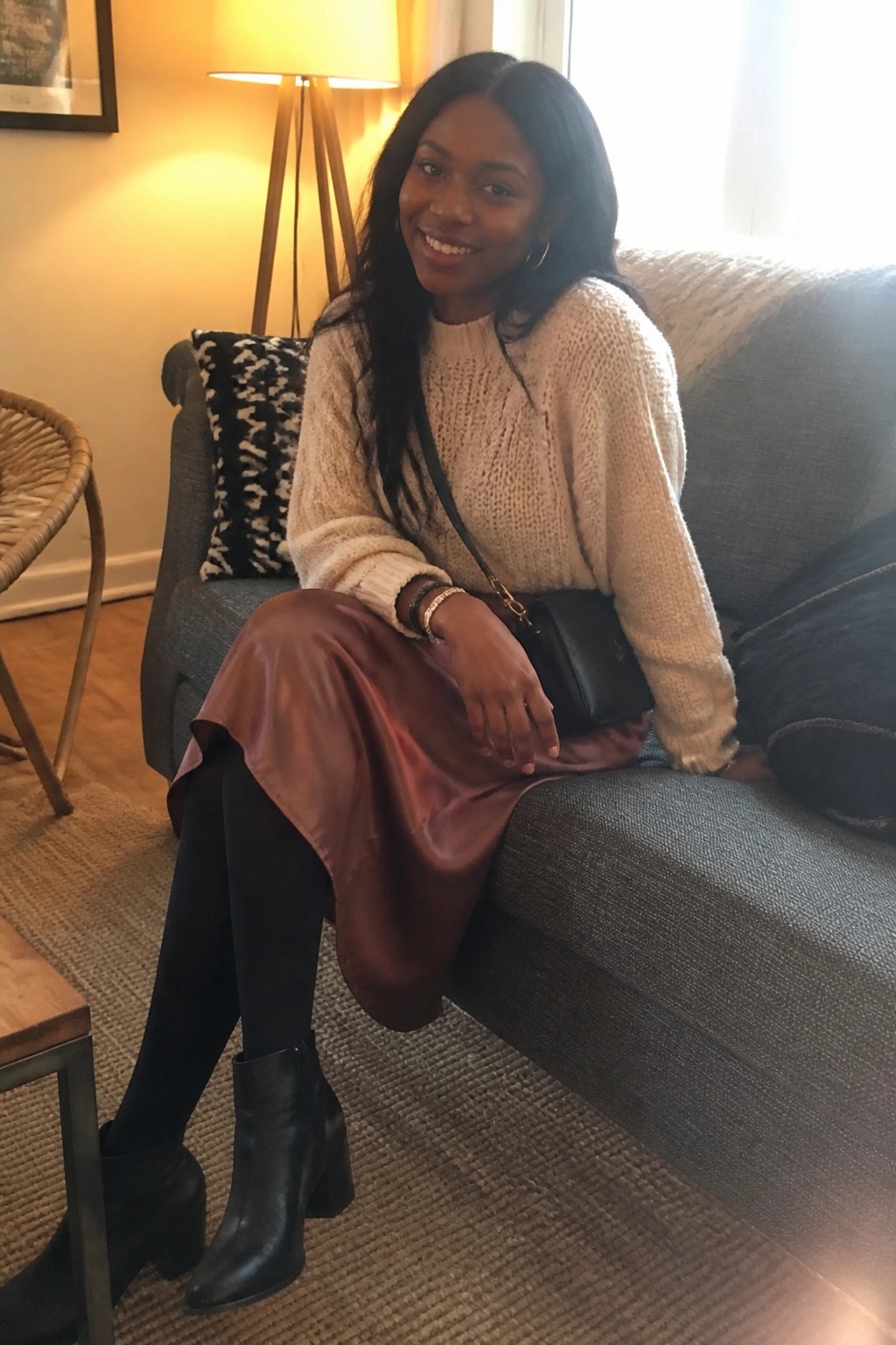 Smiling woman on gray couch in cream knit sweater, burgundy satin skirt and black boots
