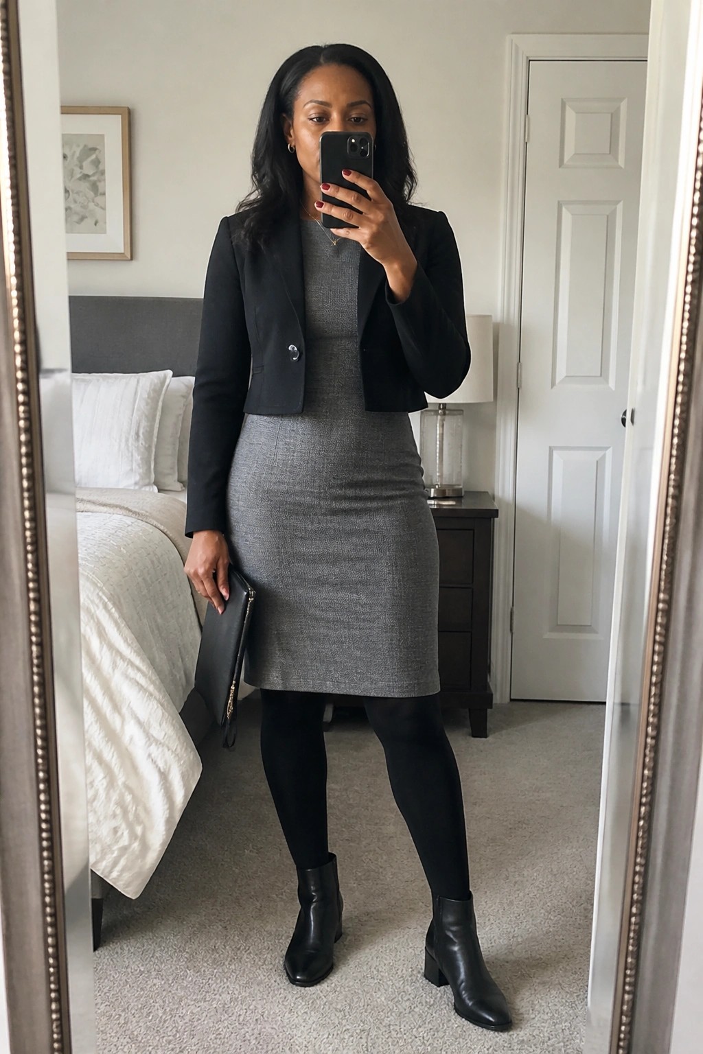 Woman in black cropped blazer and gray knit dress taking bedroom mirror selfie