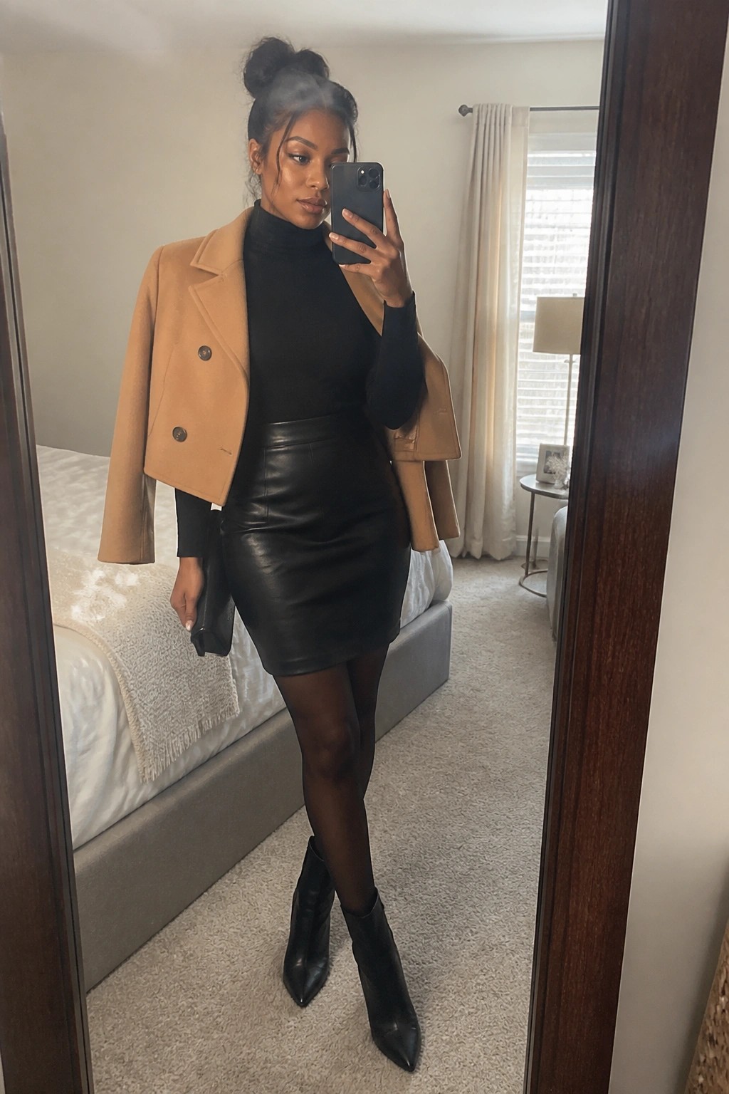 Woman in tan coat, black turtleneck, leather skirt taking mirror selfie in bedroom.