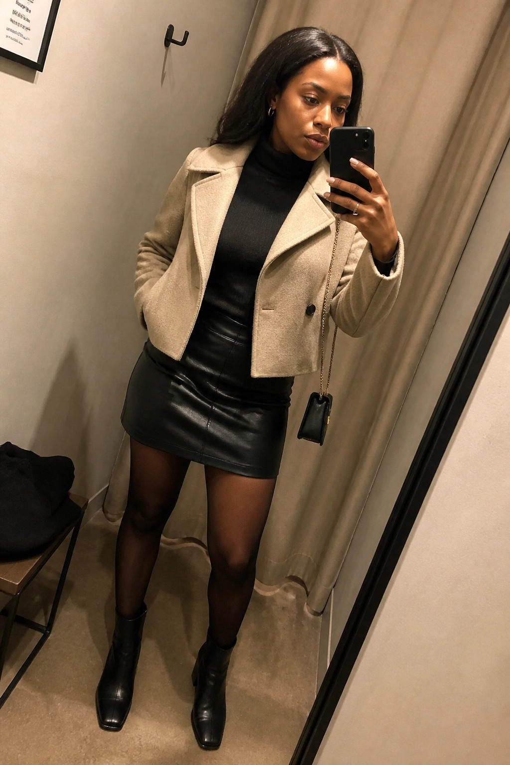 Woman wearing a beige cropped jacket over a black turtleneck with a black leather mini skirt, sheer tights, and black ankle boots while holding a phone for a mirror selfie.