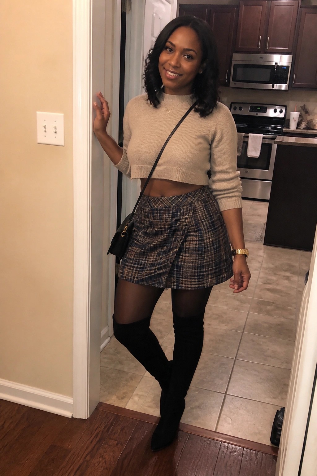 Woman in beige cropped sweater, plaid mini skirt, and black thigh-high boots posing in kitchen