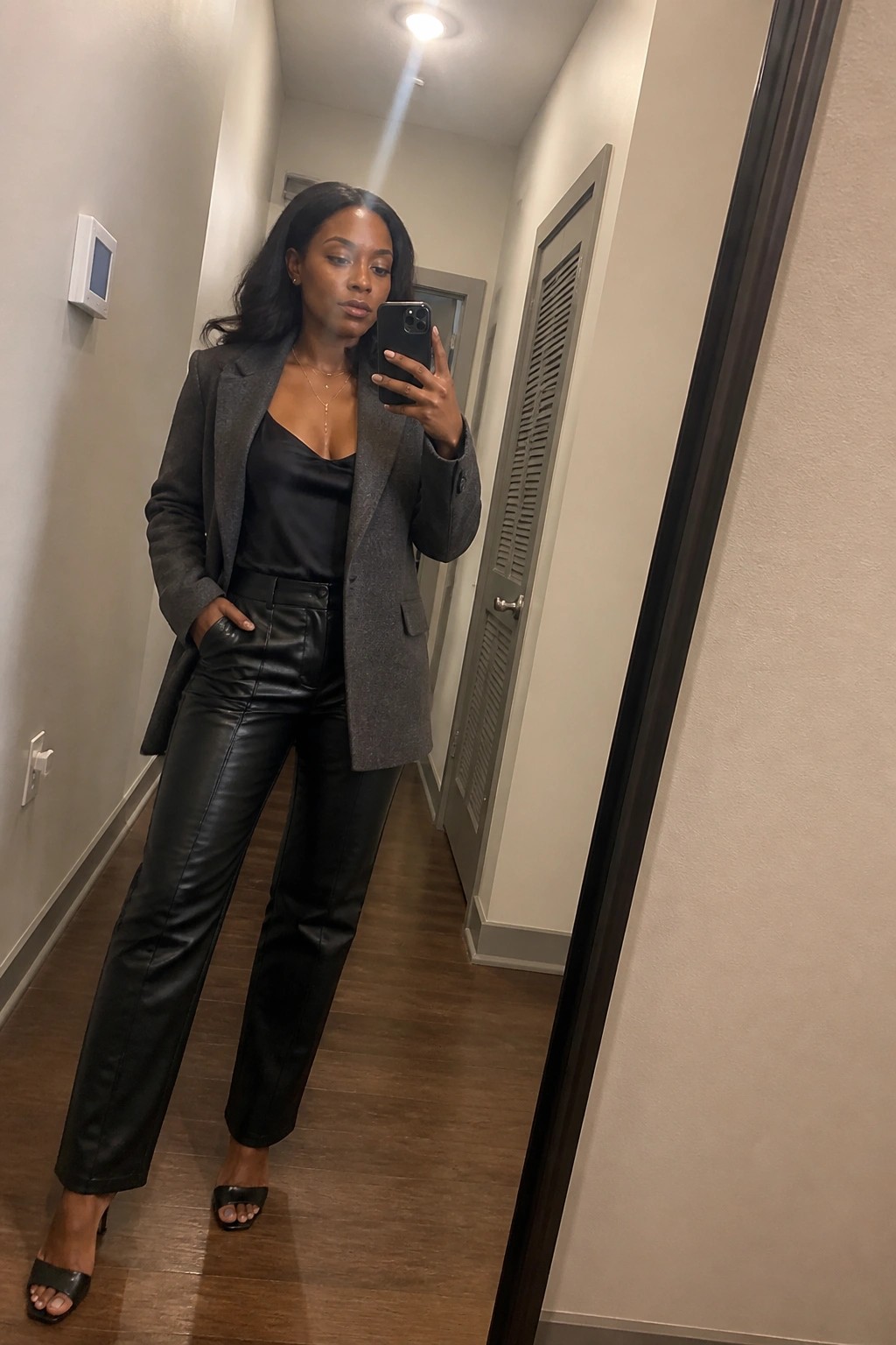 Woman in gray blazer and black leather pants takes mirror selfie in hallway.