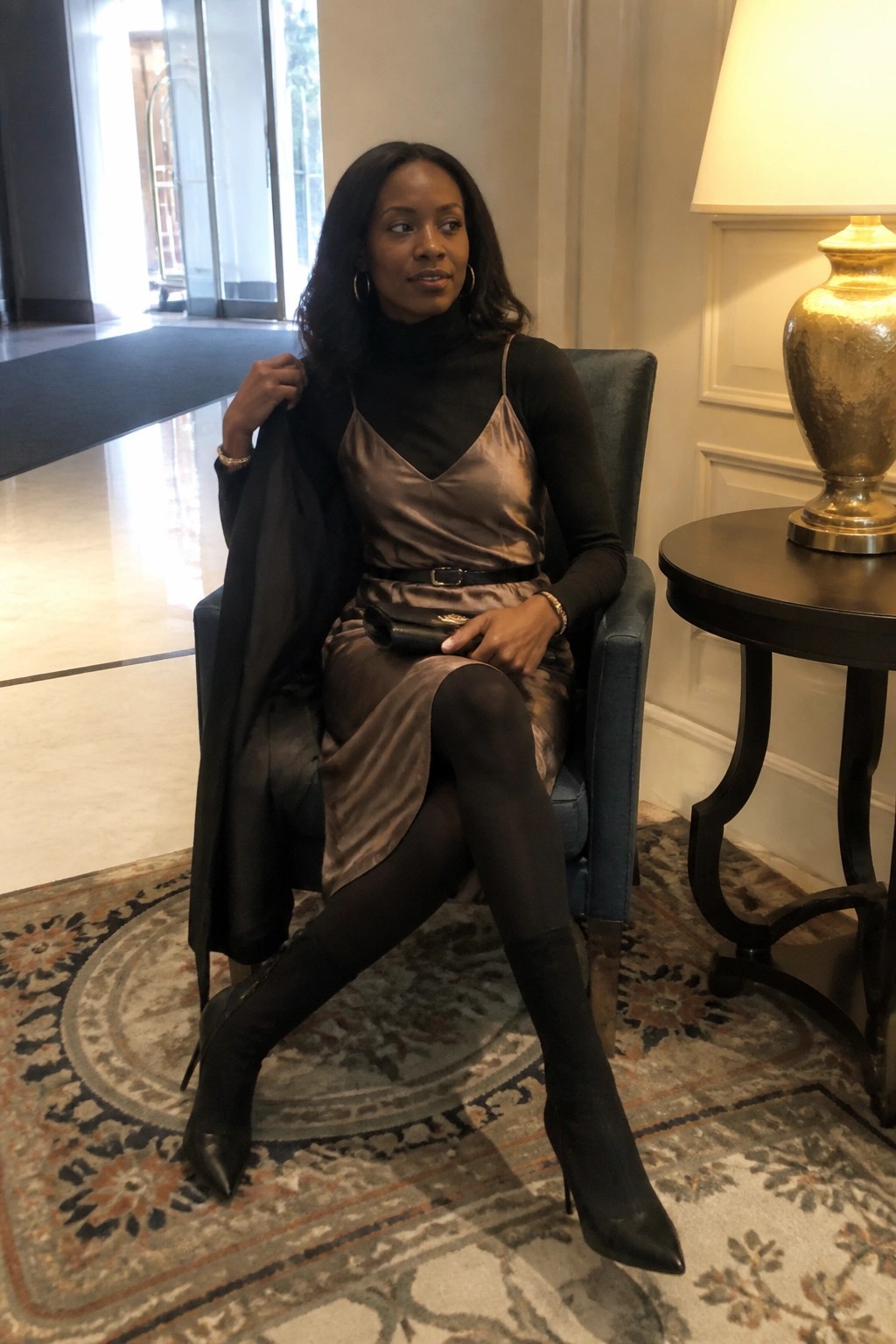 Woman in taupe satin dress and black boots holding coat in blue armchair