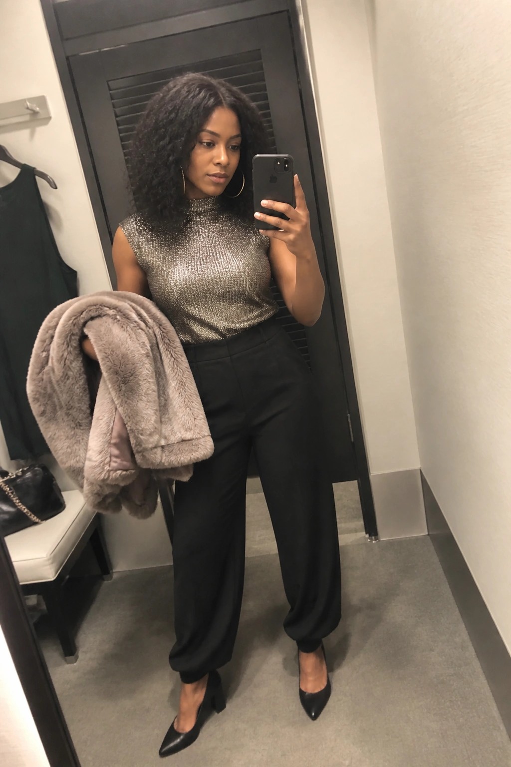 Woman in sparkly silver top and black pants holds gray fur coat in mirror selfie