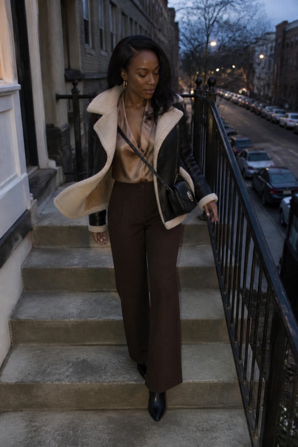 Woman in black shearling jacket and brown pants descending urban stone steps at dusk.