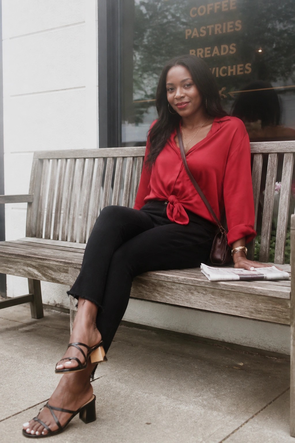 Woman in red knotted blouse and black pants seated on wooden bench outside cafe