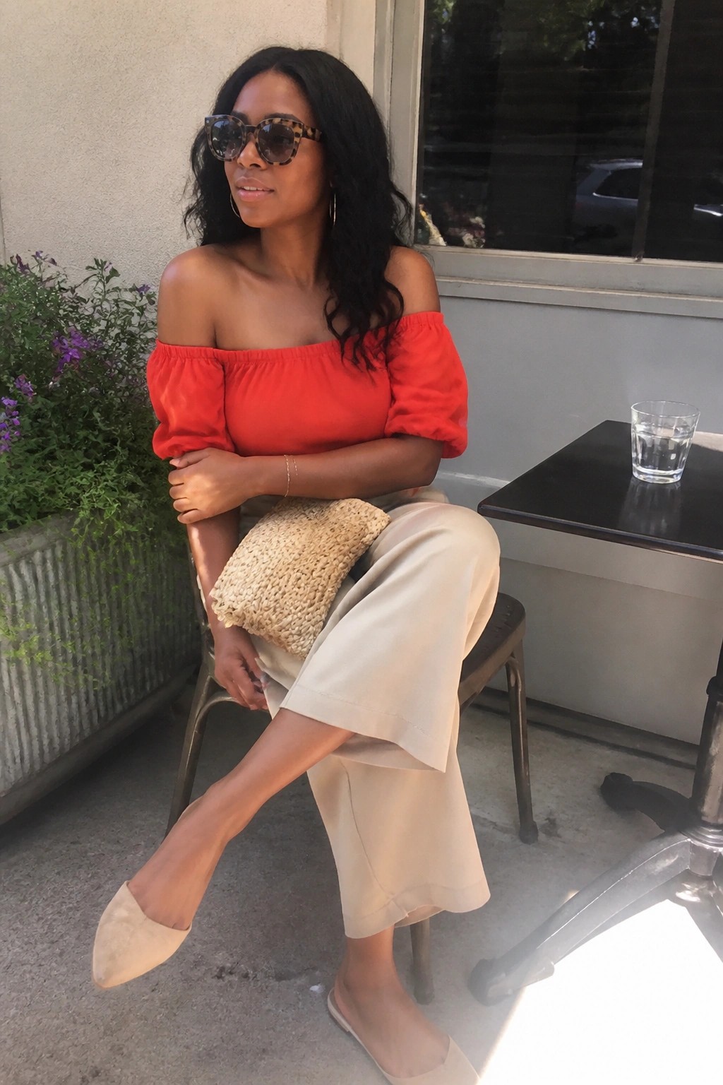 Woman in red off-shoulder top and beige pants holding woven clutch seated outdoors