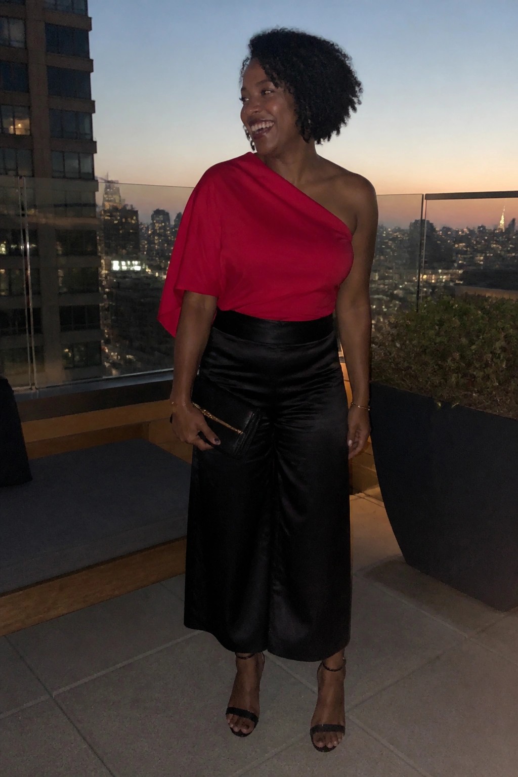 Black woman wearing a red one-shoulder asymmetric top tucked into high-waisted black satin wide-leg pants, holding a small black clutch, paired with black strappy heels and gold jewelry.
