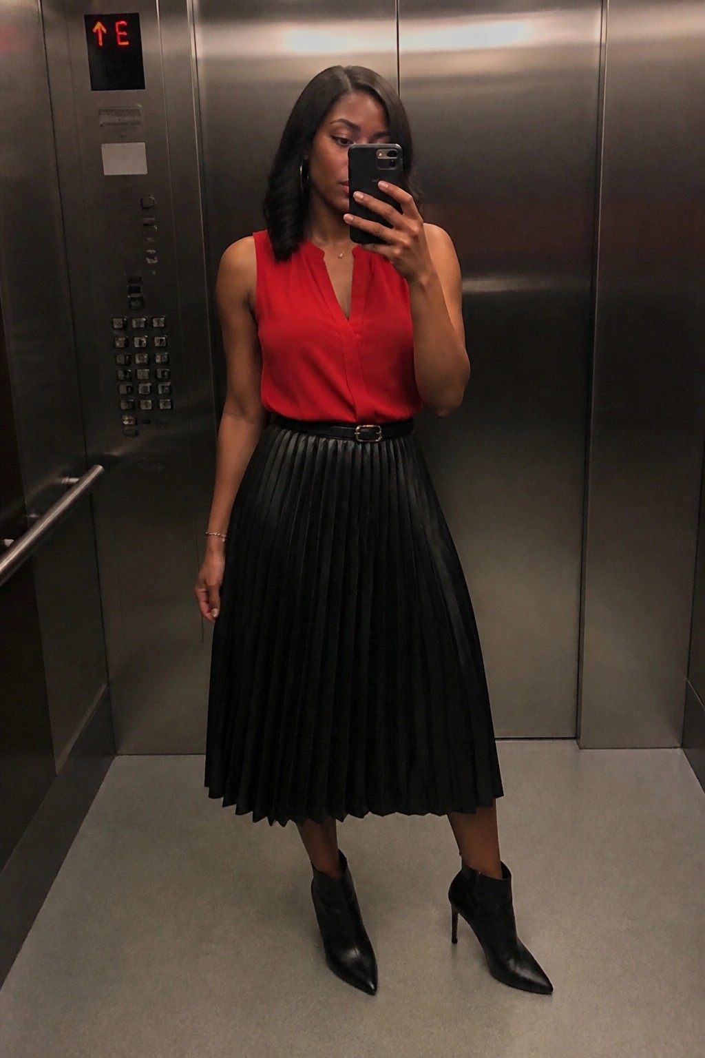 Woman in red sleeveless top and black pleated skirt taking selfie in elevator