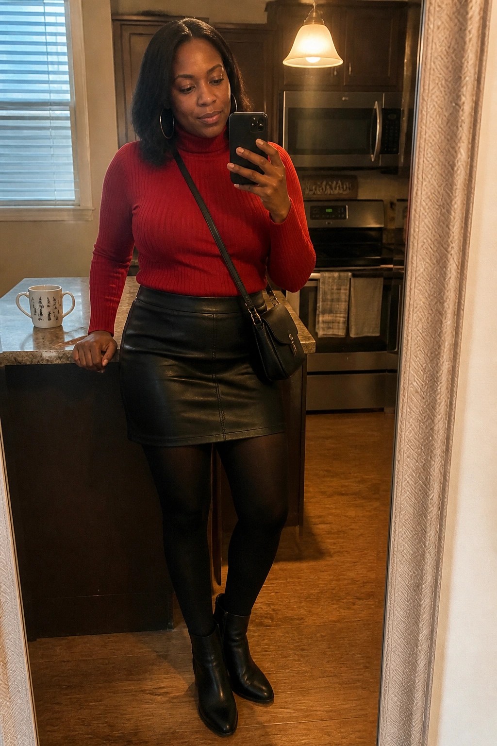 Woman wearing a fitted red ribbed turtleneck sweater tucked into a black leather mini skirt with black tights black ankle boots and a small black crossbody bag while holding a phone for a mirror selfie in a kitchen setting