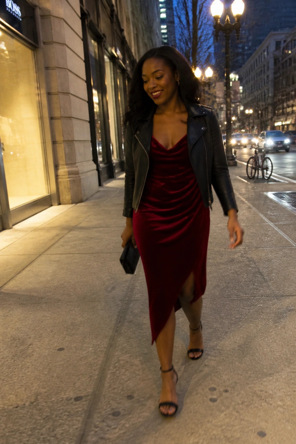 Woman in red velvet dress and black leather jacket walking on city sidewalk at night