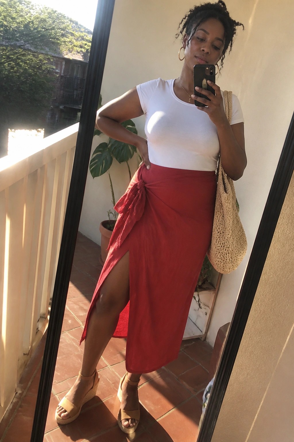 Woman in white top and red wrap skirt taking sunny balcony mirror selfie