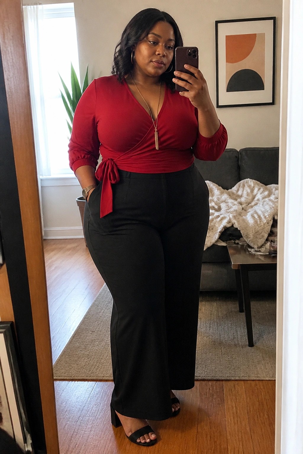 Woman in red wrap top and black pants taking mirror selfie in living room