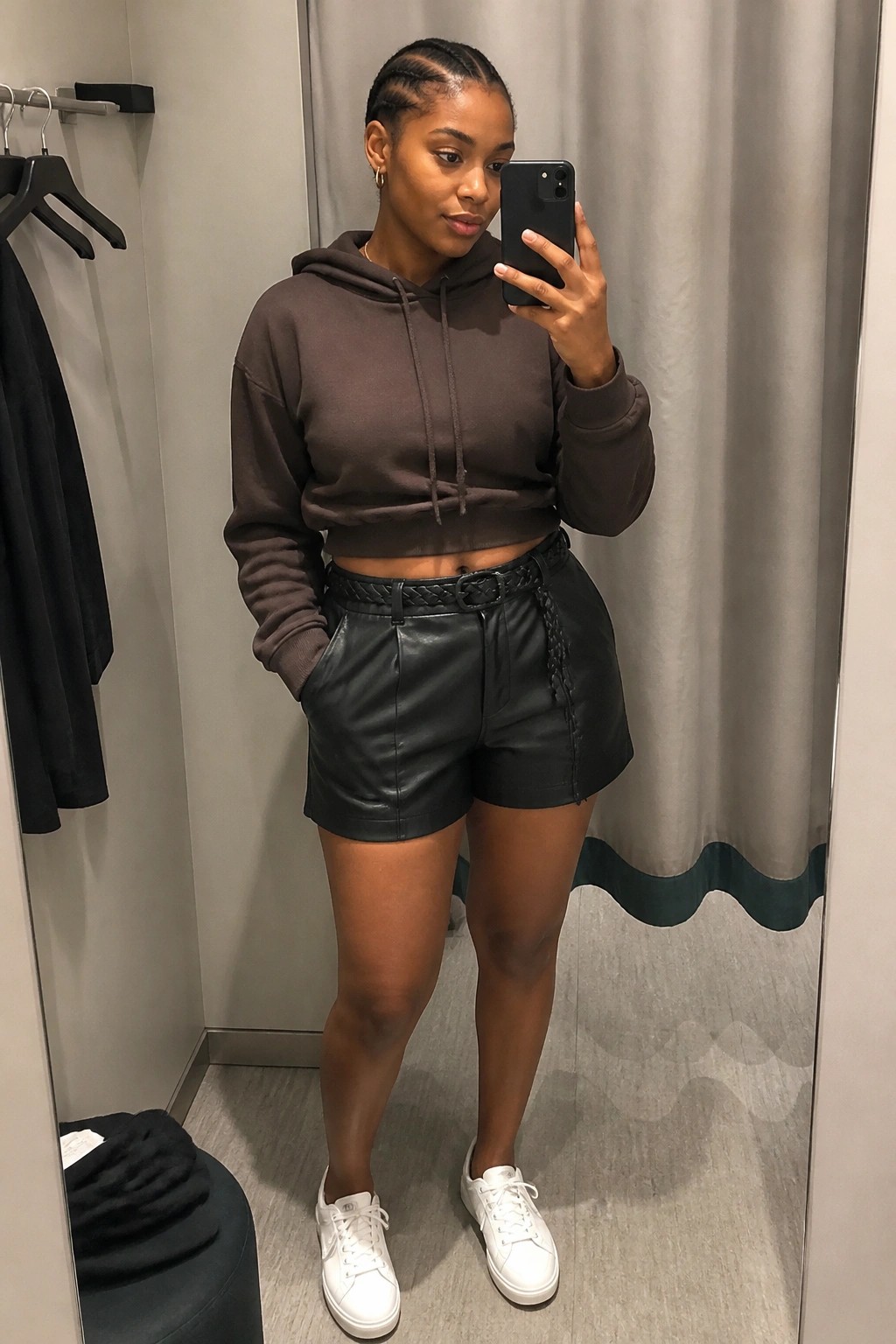 Woman in brown cropped hoodie and black leather shorts taking mirror selfie.
