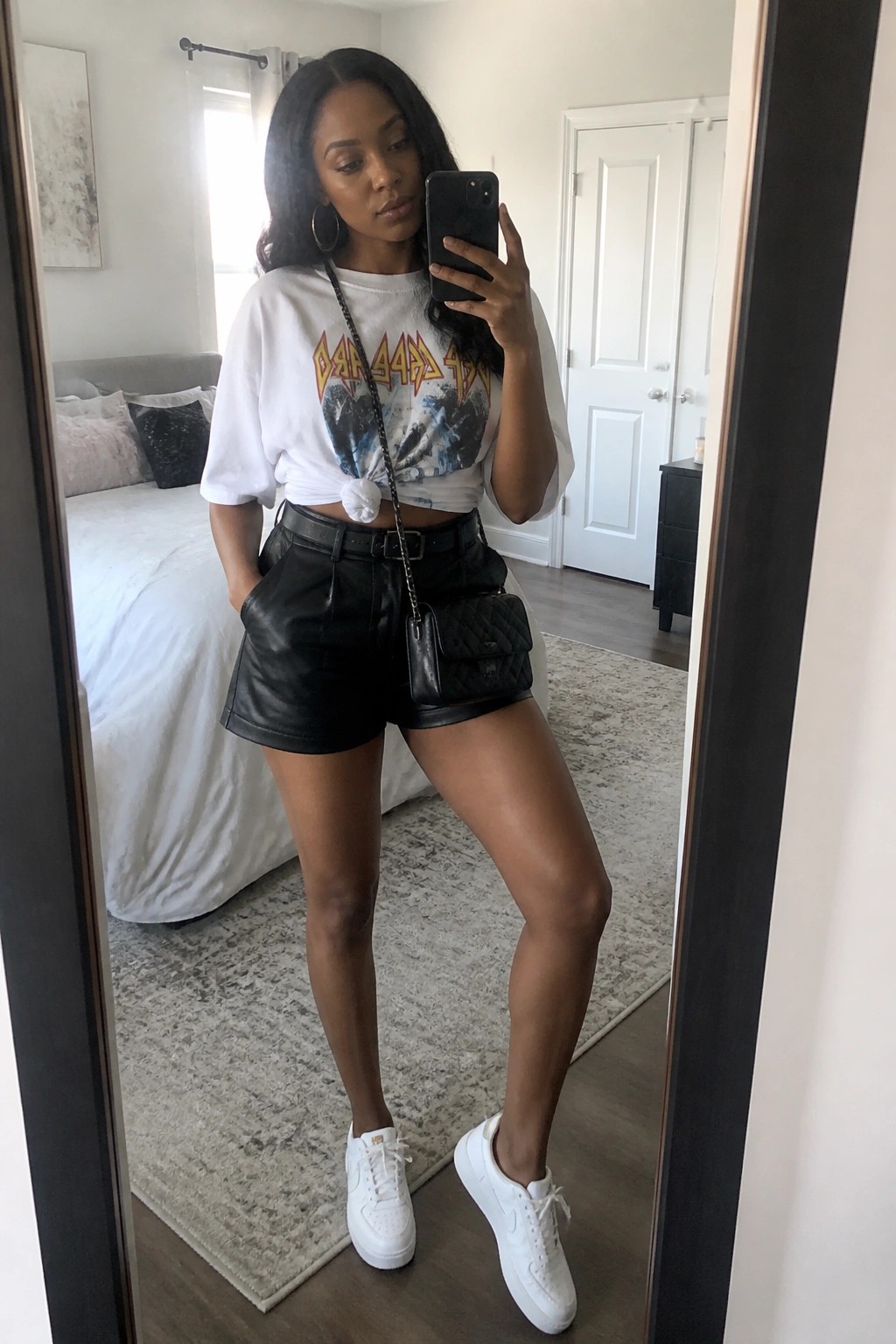 Woman in white graphic tee and black leather shorts taking bedroom mirror selfie