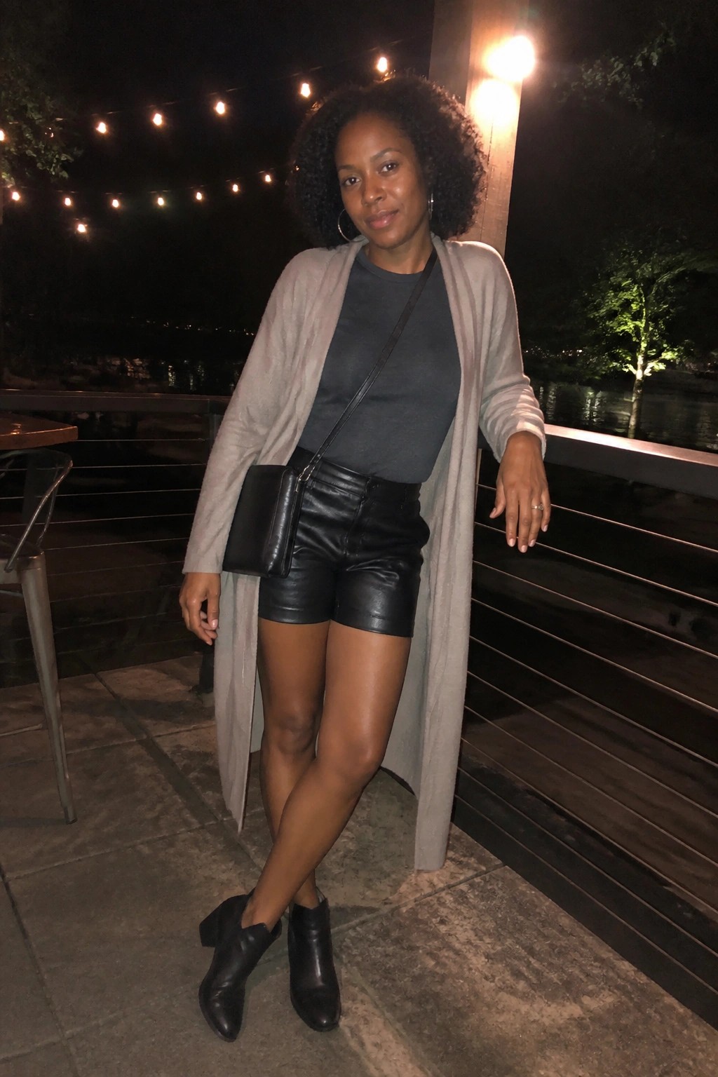 Woman wearing black leather shorts, gray crewneck top, long beige cardigan, black crossbody bag, and black ankle boots standing on an outdoor deck at night.