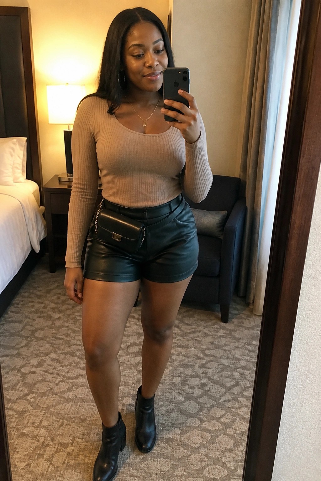 Woman in beige ribbed top and black leather shorts taking mirror selfie in hotel room