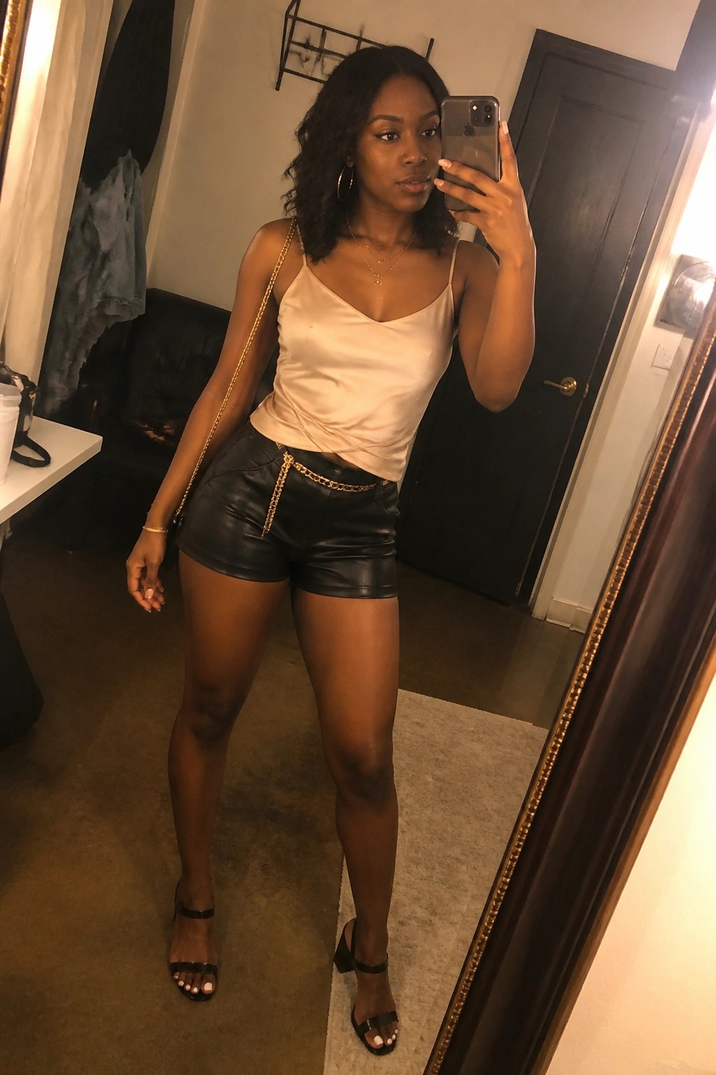 Woman in beige satin camisole and black leather shorts with gold chains taking mirror selfie.