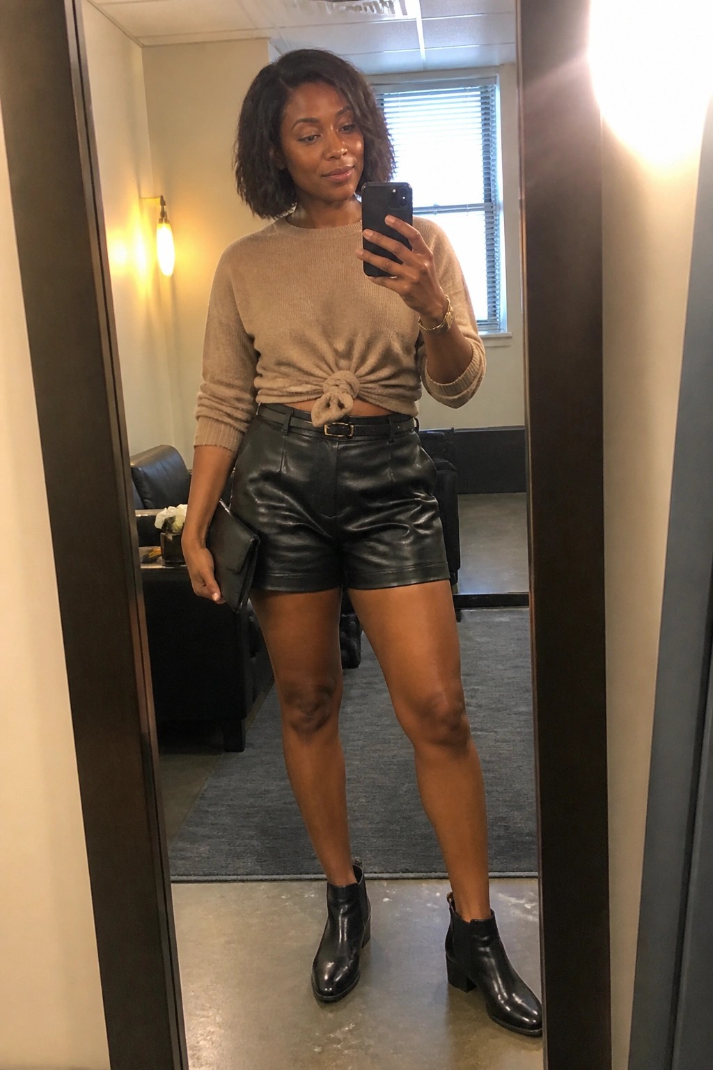 A woman wearing a beige knit sweater tied at the waist with black leather shorts, black belt, black ankle boots, and holding a black clutch while taking a mirror selfie.