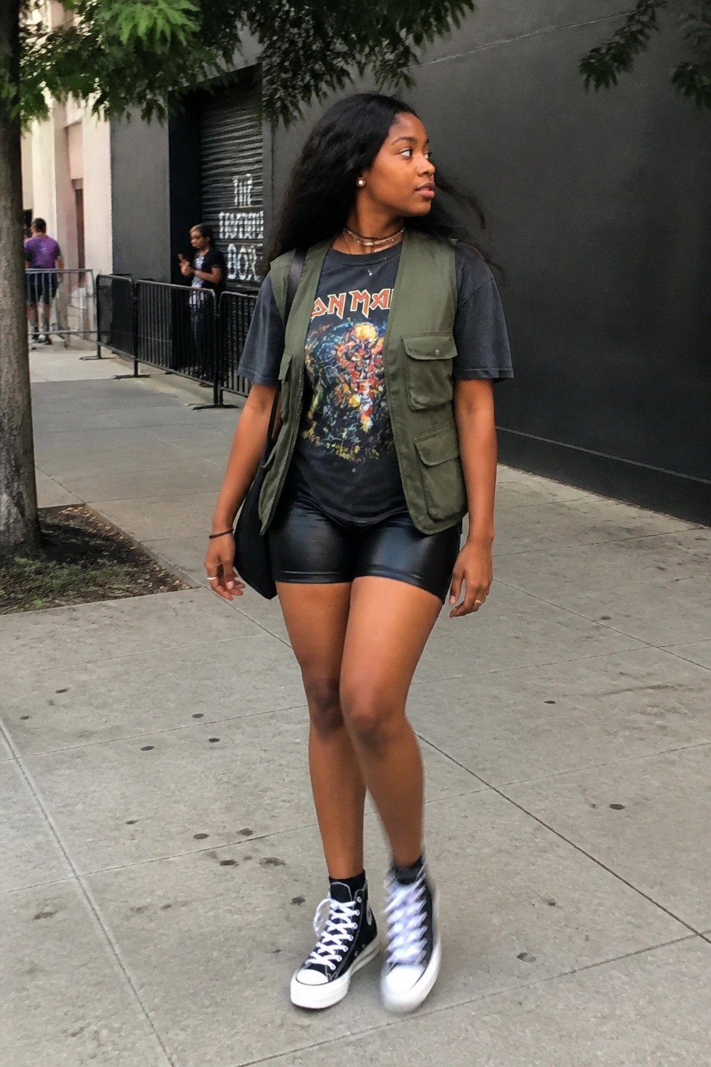 Woman in green utility vest over Iron Maiden tee with black shorts on sidewalk.