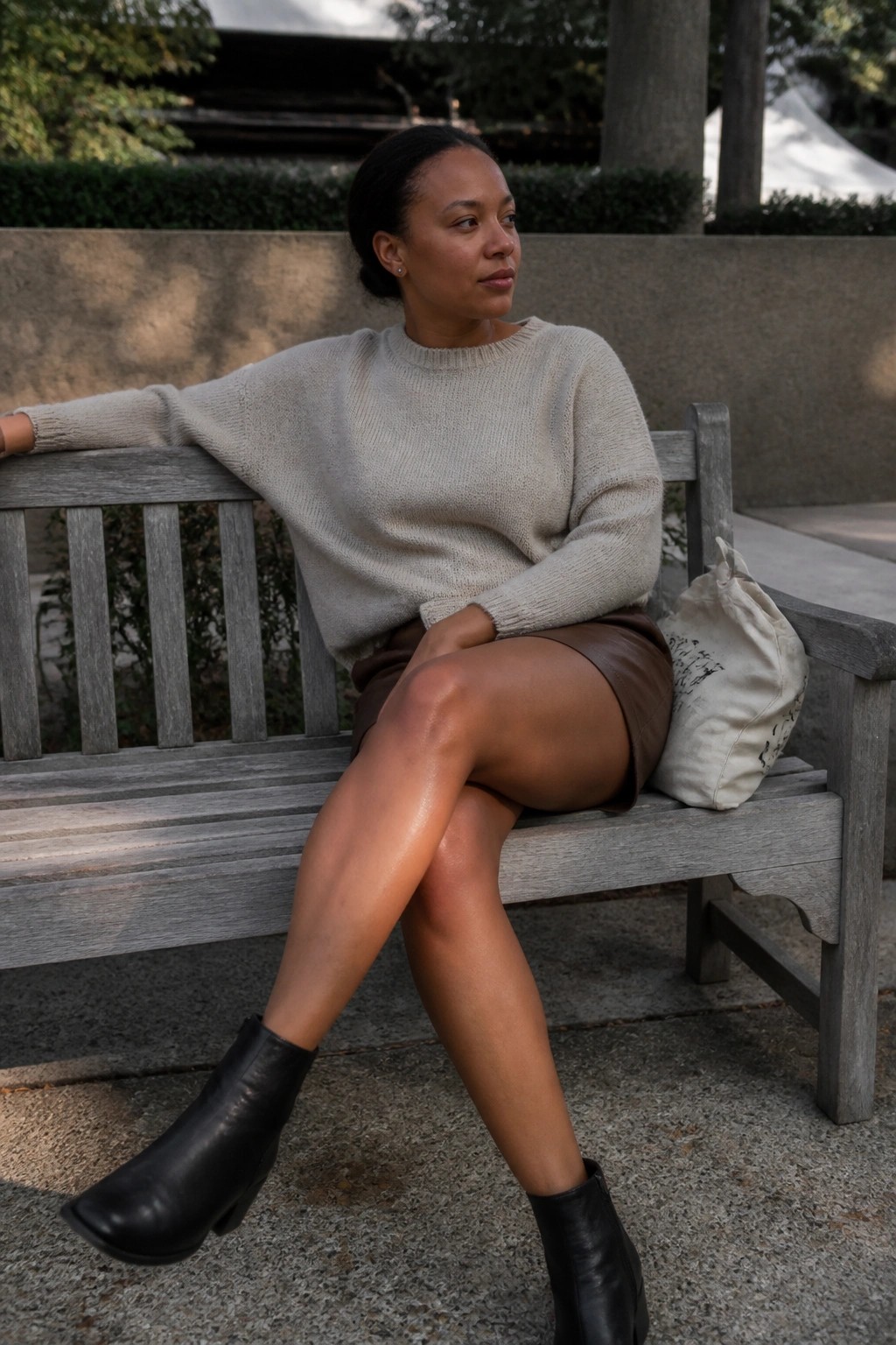 Woman in oversized beige knit sweater, brown leather shorts, and black ankle boots seated on a bench with a canvas tote nearby.