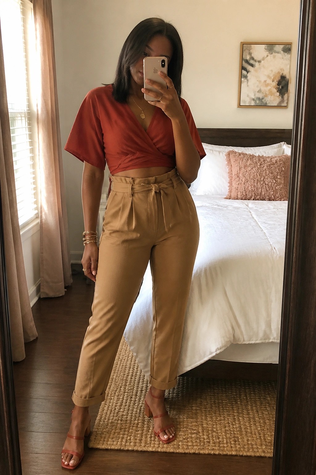 Woman in rust wrap top and tan belted pants taking bedroom mirror selfie.