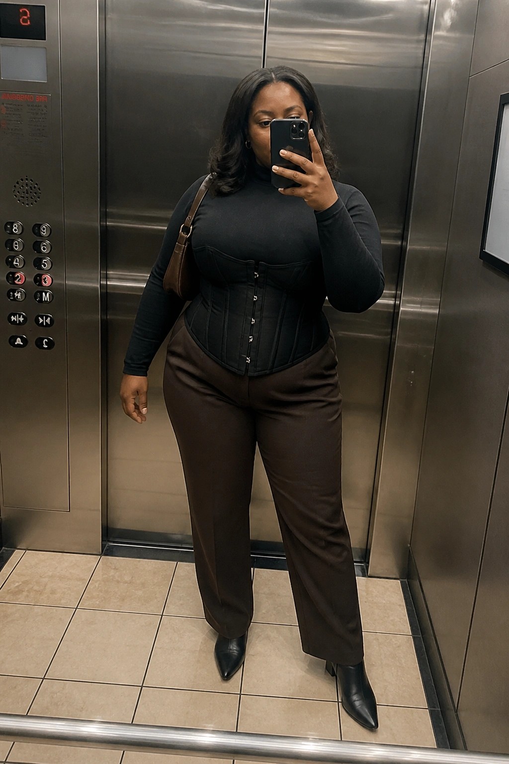 Plus size Black woman in elevator wearing black corset over black turtleneck, brown wide leg pants, black pointed boots, and brown shoulder bag while taking a mirror selfie.