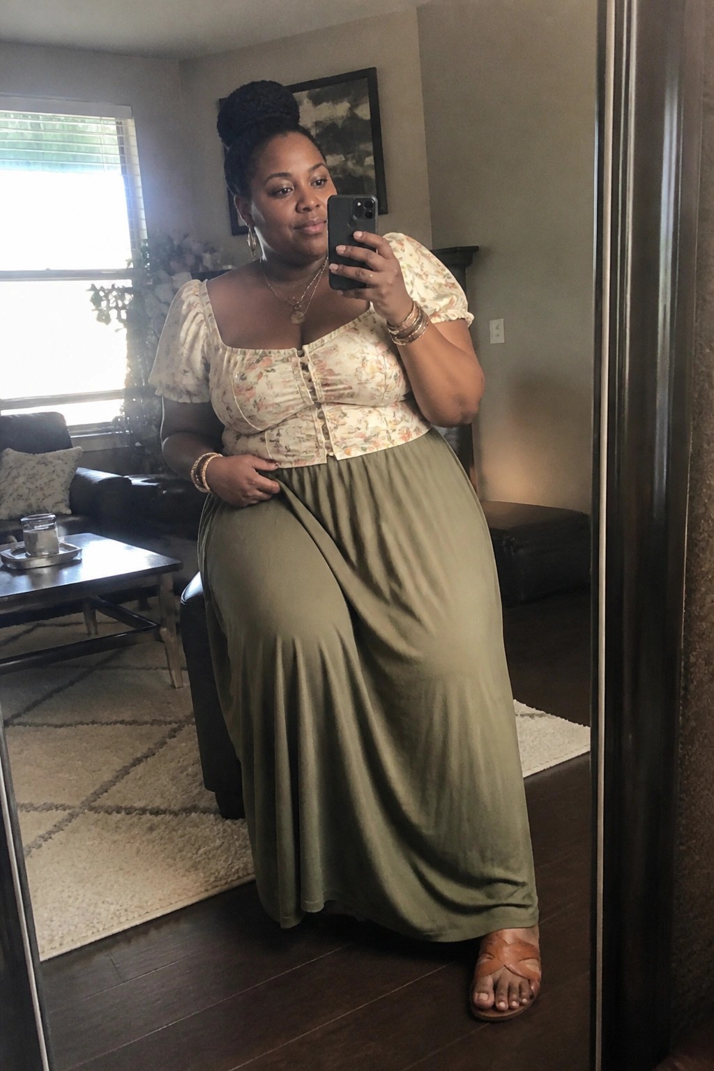 Woman in floral top and olive green skirt taking mirror selfie in living room