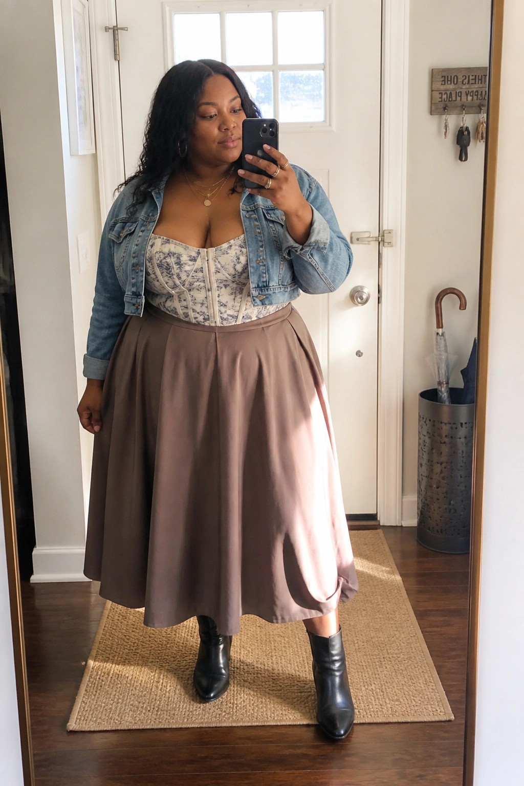 Plus-size woman in denim jacket, floral corset, taupe midi skirt, and black boots taking selfie.