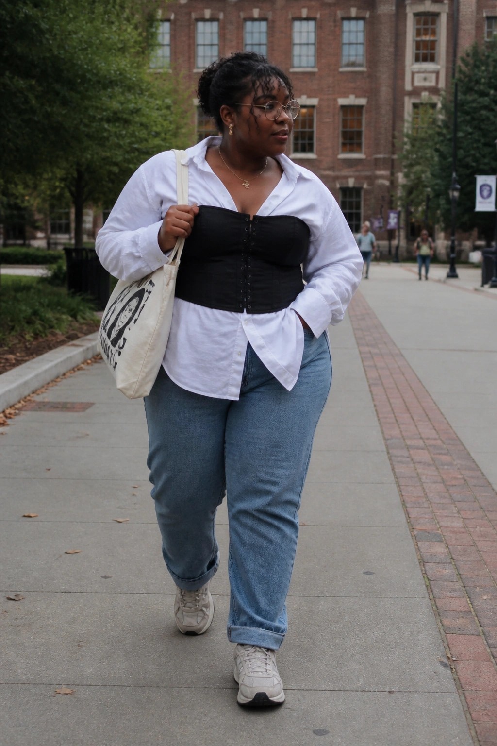 Woman in white shirt over black corset and blue jeans walks with tote bag on path.